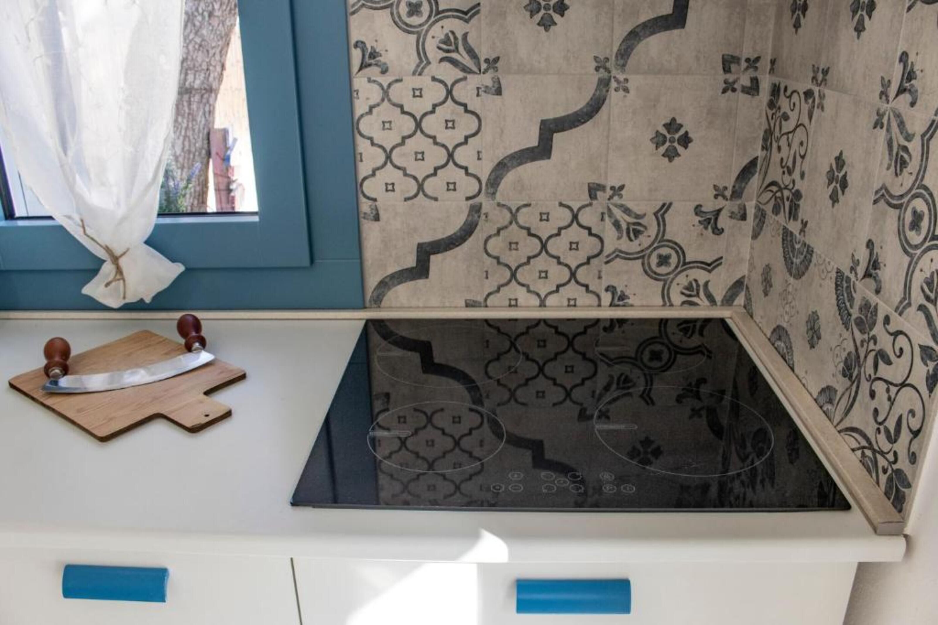 A kitchen with a patterned backsplash, a window with a curtain, and a wooden cutting board with a knife.