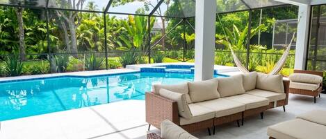 A modern outdoor pool area with a glass enclosure, wicker furniture, and lush greenery in the background.