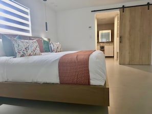 A modern bedroom with a bed, pillows, a wooden wardrobe, and a kitchen area visible through an open door.