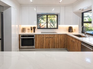 A modern kitchen with white cabinets, wooden lower cabinets, a built-in oven, a sink, and a window with a view of greenery. - Coral Springs Oasis Retreat (Coral Springs)
