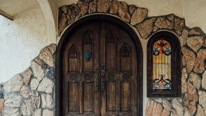 Uma grande porta dupla de madeira, com arco de pedra acima e uma janela de vitrais ao lado direito.