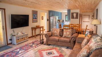 A living room with a sofa, a television, a kitchen in the background, and a patterned rug.