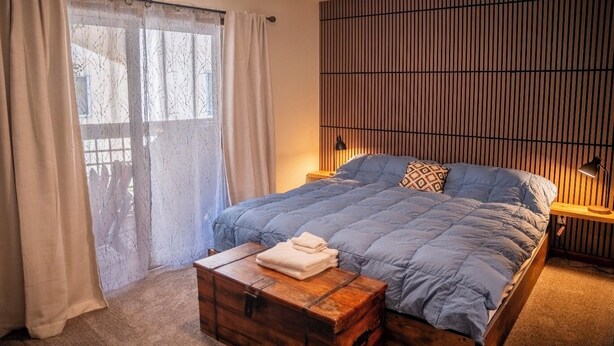 Ein Schlafzimmer mit einem Bett, einem Nachttisch, einer Holzkiste und einem Fenster mit Vorhängen.