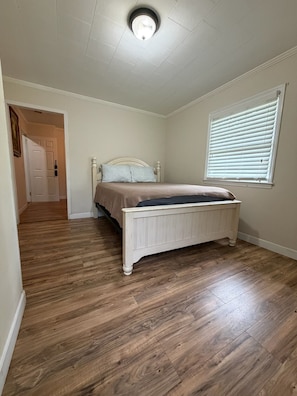 A bedroom with a wooden floor, a white bed, a window with blinds, and a door leading to another room.