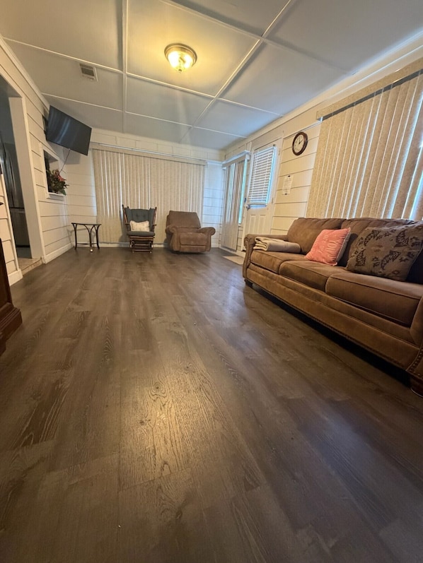 A living room with a wooden floor, a brown sofa, two armchairs, and a TV mounted on the wall.