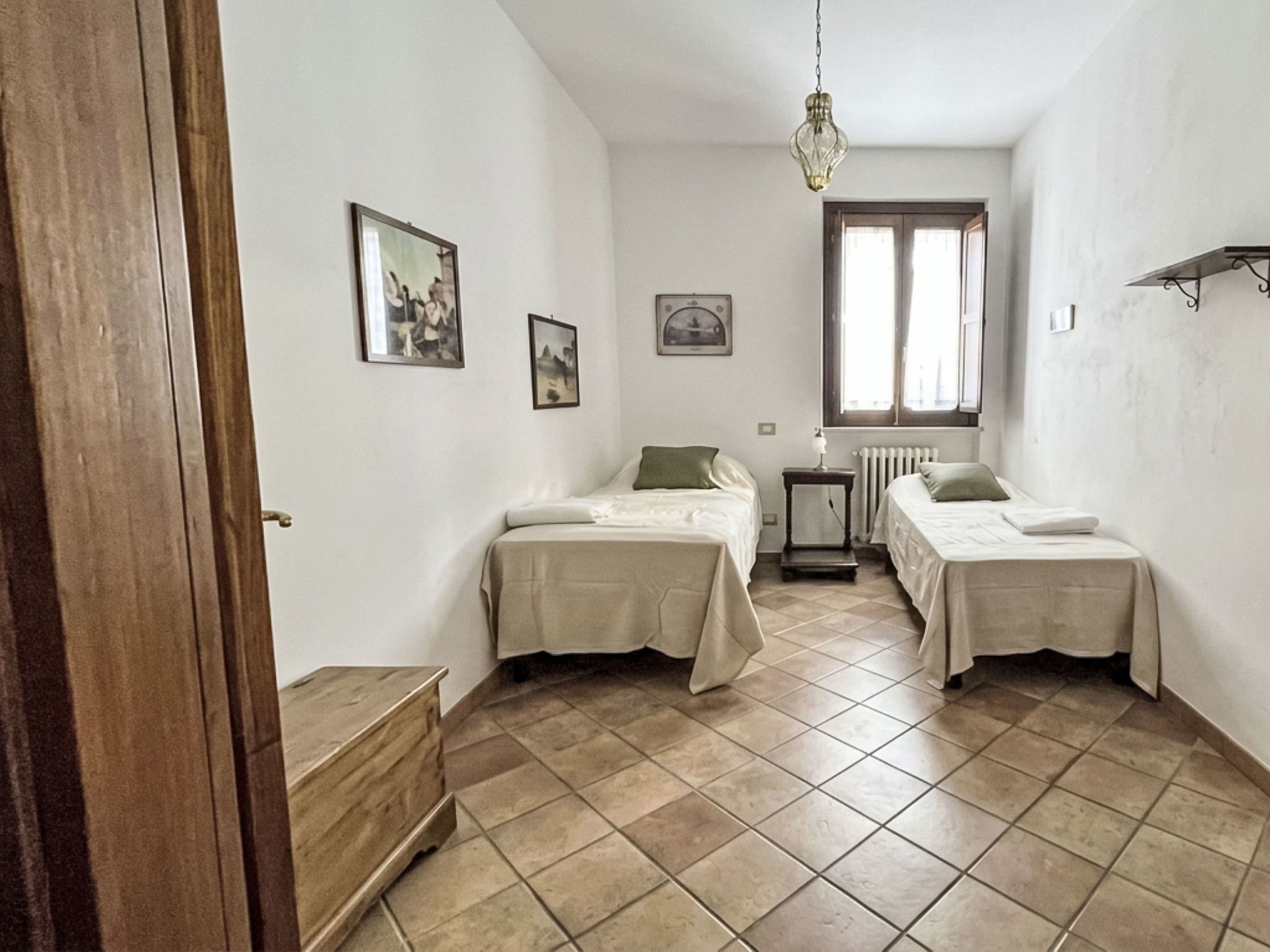 A room with two beds, a radiator, a window, and a hanging light fixture.