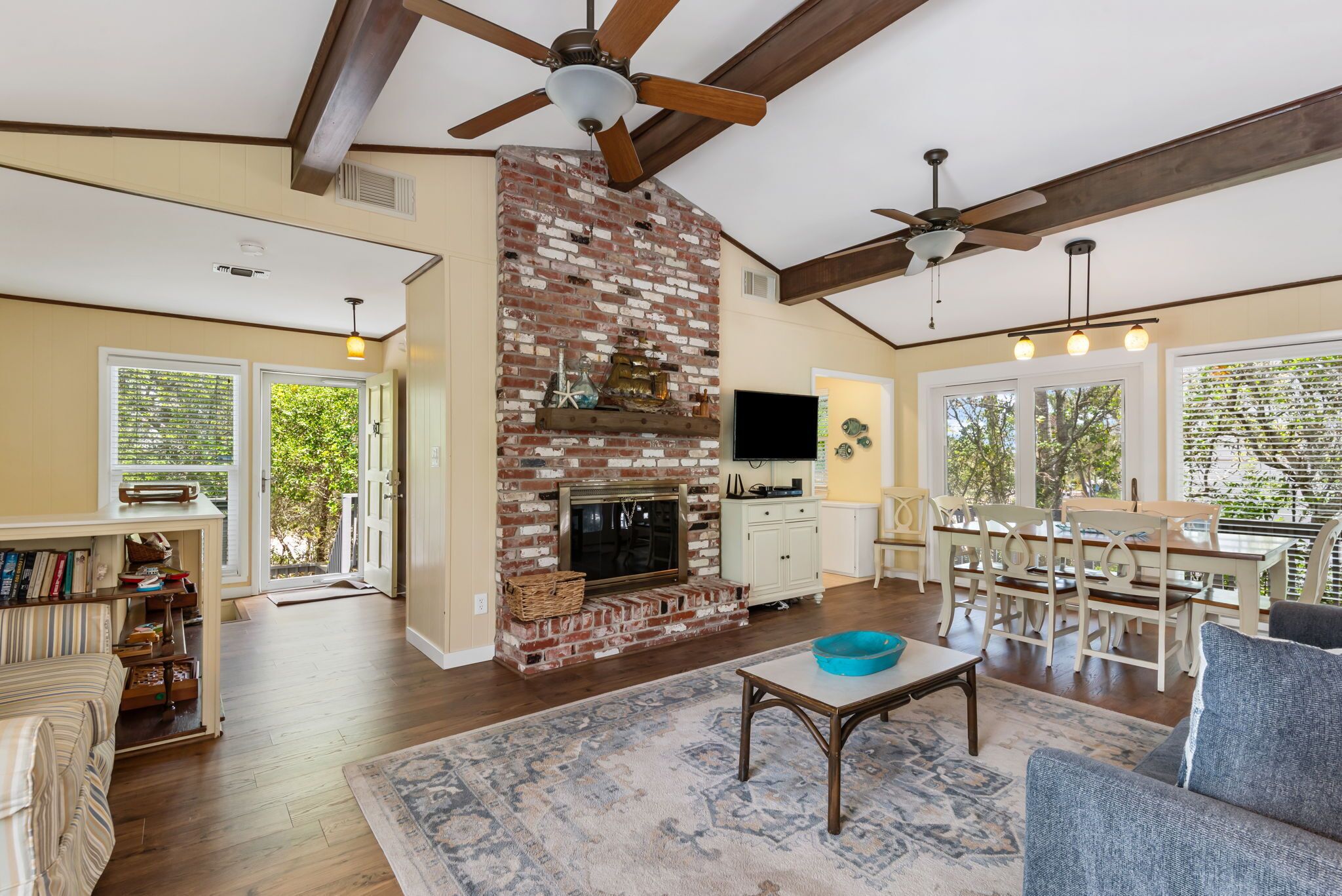 A spacious living room with a brick fireplace, ceiling fans, a dining area, and a large window.