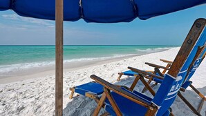 Ein Strand mit einem blauen Sonnenschirm, Holzliegen und einem sandigen Ufer.