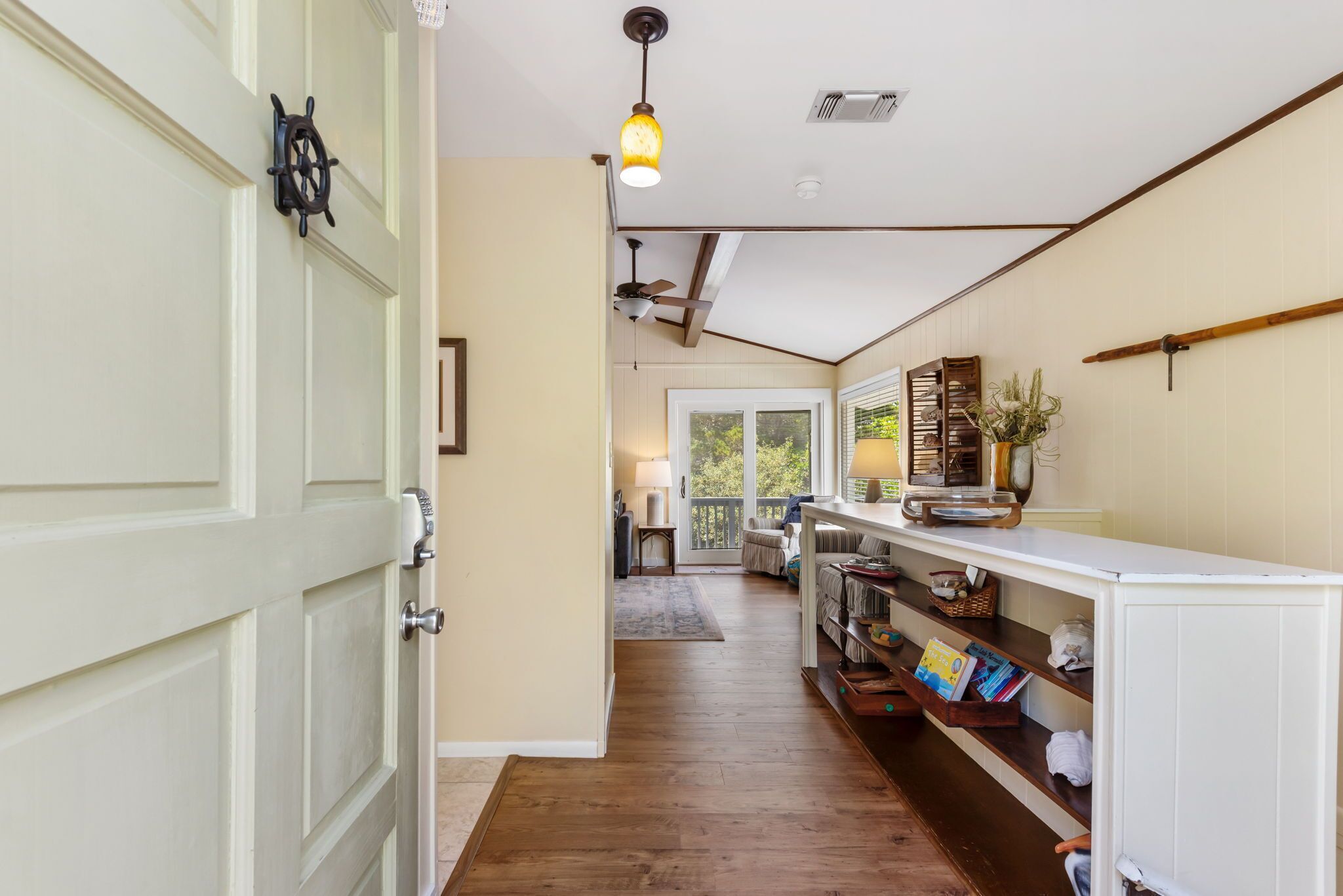 A hallway with wooden flooring, a white door with a ship's wheel, a ceiling fan, and a white console table with books and decorative items.