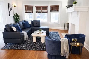 Een woonkamer met een modulaire bank, een ronde salontafel, een blauwe fauteuil en een open haard.
