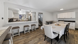 Una cocina moderna con muebles blancos, un comedor con mesa y sillas de madera, y una escalera que conduce a otro nivel.