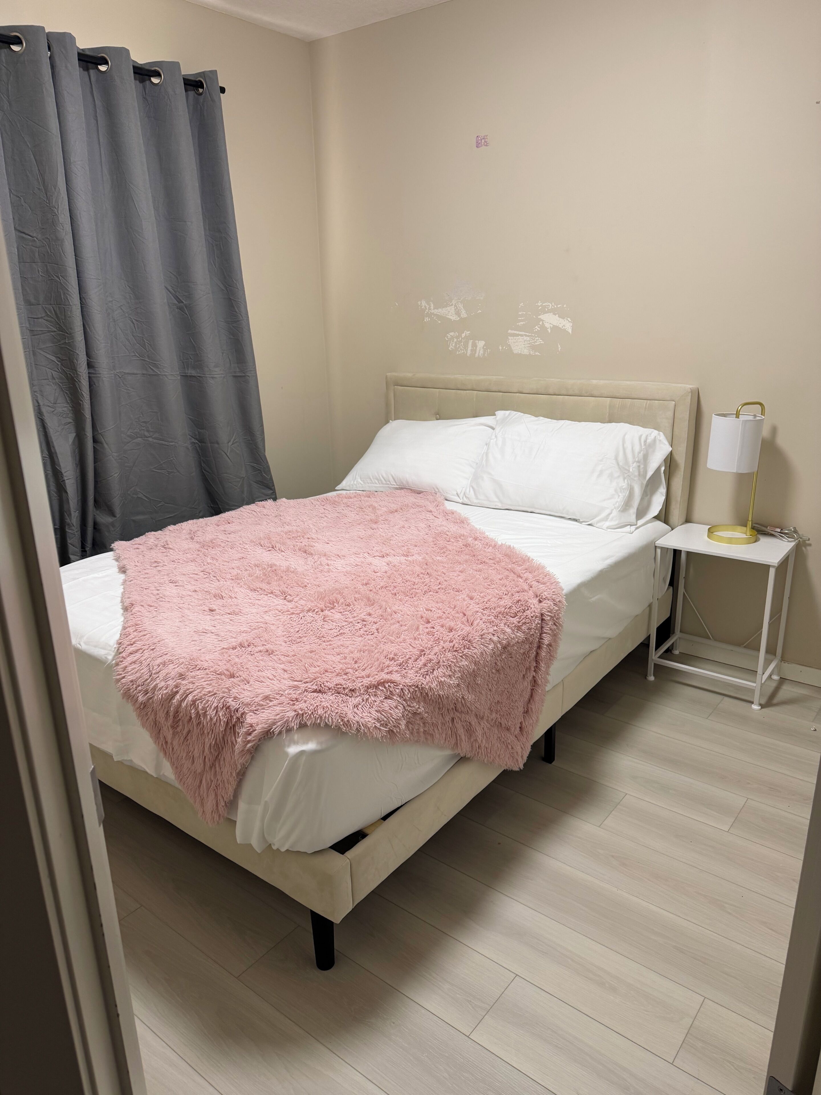 Un letto con una coperta rosa soffice, un cuscino bianco e una tenda grigia.