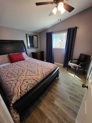 A bedroom with a bed, a ceiling fan, a dresser, a chair, a window with curtains, and a door.
