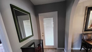 A hallway with a dark wooden floor, a dresser, a mirror, and a door with blinds.