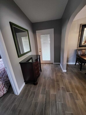 A hallway with a dark wooden floor, a dresser, a mirror, and a door with blinds.