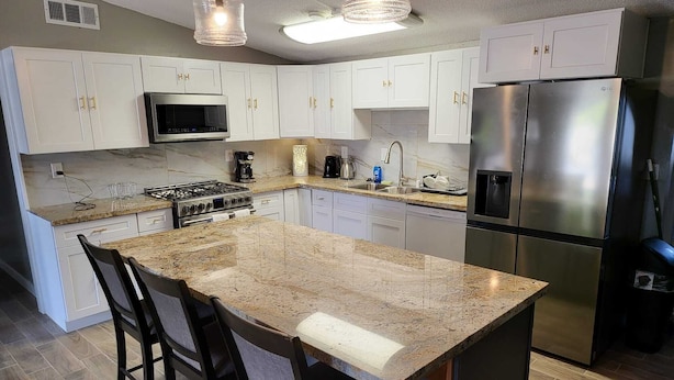 A kitchen with white cabinets, a stainless steel refrigerator, a microwave, a stove, and a large granite island with bar stools.
