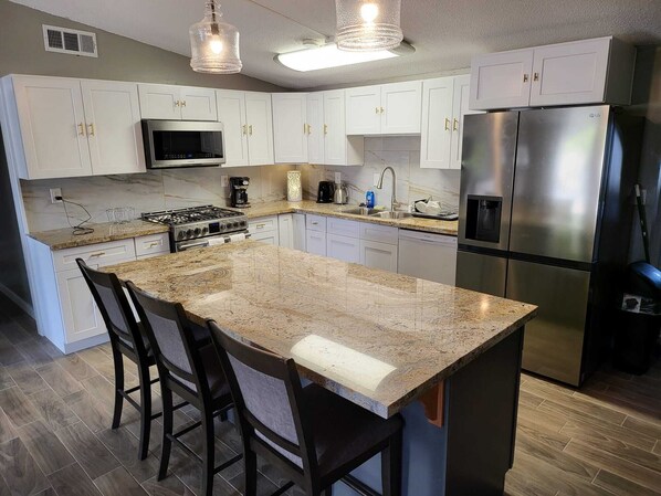 A kitchen with white cabinets, a stainless steel refrigerator, a microwave, a stove, and a large granite island with bar stools.