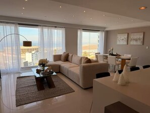 Una sala moderna con sofá, mesa de comedor y ventanas amplias con cortinas.