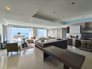 A modern living room with a large island, a kitchen with dark cabinets, and a view of the city. - Sky Penthouse with Panoramic Ocean View & Jacuzzi (Tijuana)