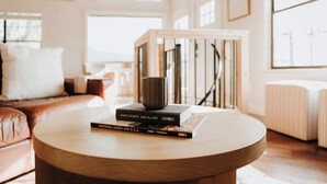 Un tavolo rotondo in legno con libri e una tazza, un divano in pelle e un termosifone bianco in una stanza luminosa con ampie finestre.