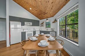 Une cuisine moderne avec des appareils électroménagers blancs, une table à manger en bois dressée avec de l’assiette blanche, et une vue sur la verdure grâce à de grandes fenêtres.