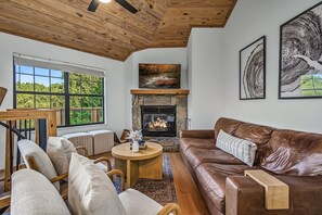 A cozy living room with a fireplace, a large window, and a leather couch. - Mountain Vista: Modern Cabin with Mt. LeConte View & Hot Tub (Gatlinburg)
