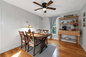 A dining area with a wooden table and chairs, a ceiling fan, a kitchen area with a wooden cabinet, and a window with curtains. - Canvas & Coast DT TC, Games + Fire Pit | Pets Ok (Traverse City)