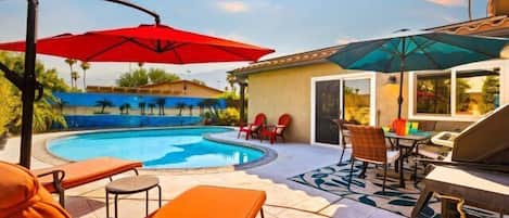 Une piscine avec des chaises longues et des parasols, un patio avec une table et des chaises, et un bâtiment doté d’un toit en tuiles.