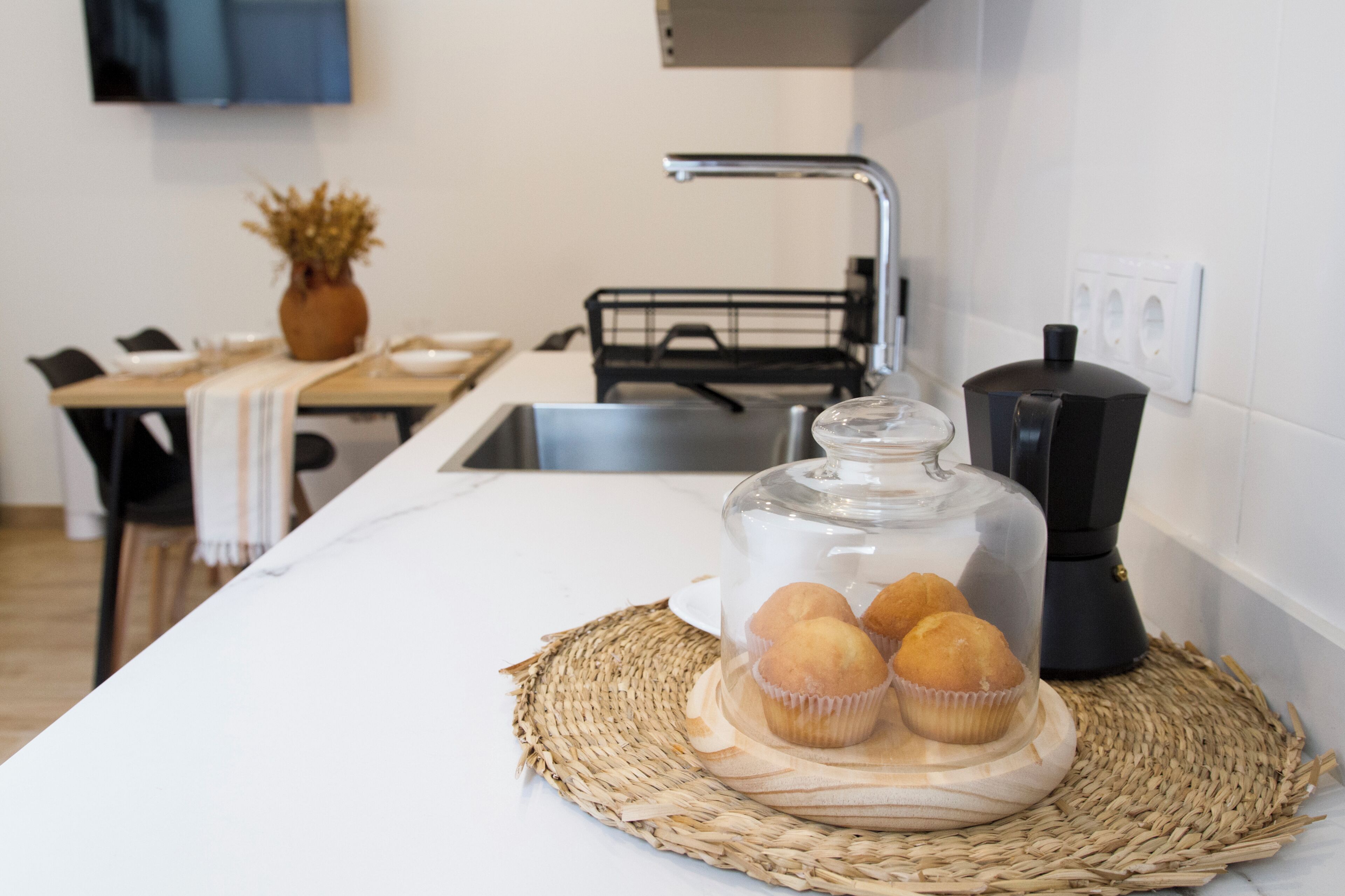 Une cuisine avec un récipient en verre transparent contenant des muffins, posé sur un set de table en fibres tressées, une machine à café et un coin repas avec des chaises et un vase de fleurs séchées.