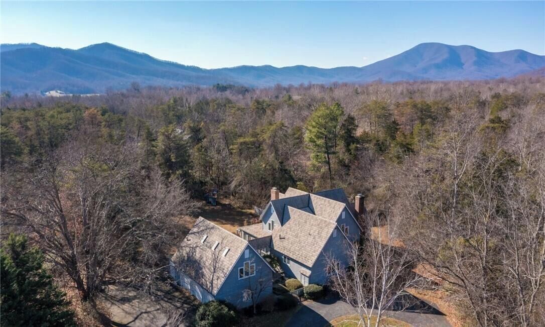 A large house with a steep roof, surrounded by trees and mountains in the background.