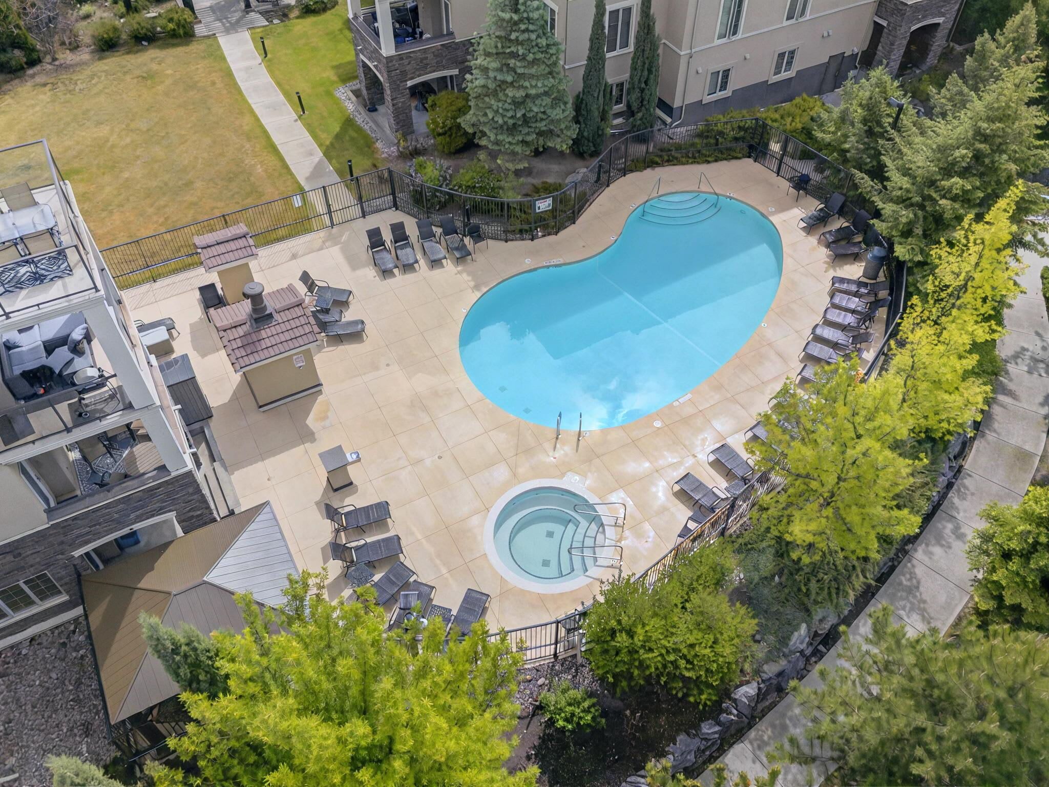 A swimming pool with lounge chairs and a hot tub, surrounded by trees and a building.