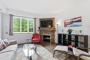 A living room with a fireplace, a TV mounted on the wall, a red chair, a glass coffee table, and a bookshelf. - Pinnacle Pointe | 2 Bed Golf Getaway (Kelowna)