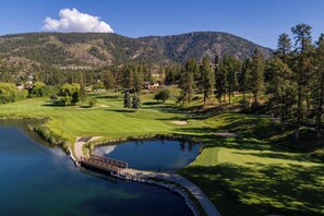 A golf course with a pond, a wooden bridge, and a mountain backdrop. - Pinnacle Pointe | 2 Bed Golf Getaway (Kelowna)