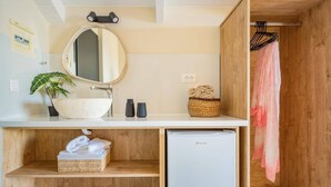 Un bagno compatto con uno specchio rotondo, un lavandino bianco, un piccolo frigorifero, una pianta e un armadietto in legno.