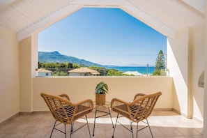 Ein Balkon mit zwei Korbstühlen und einem Blumentopf, mit Blick auf eine Küstenlandschaft mit Häusern und einem Berg.