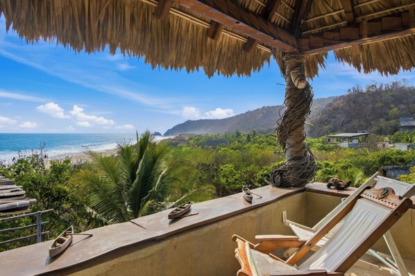 Un balcón con vista a la playa, una palmera y una estructura de techo de paja.