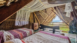 Une chambre confortable, aménagée comme une tente avec un toit de chaume, comprenant un lit, un ventilateur et offrant une vue sur l’extérieur.