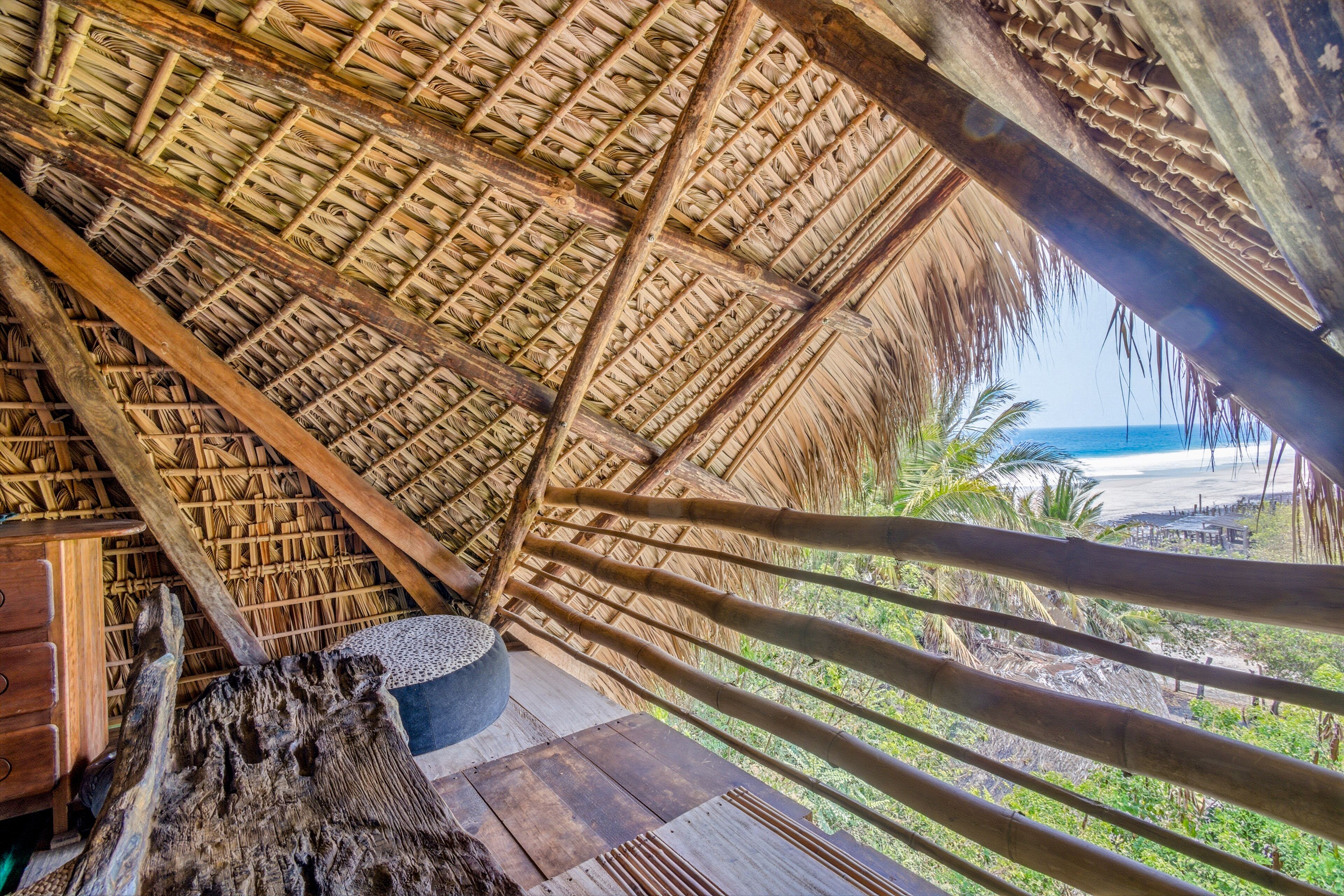 Une construction à toit de chaume, dotée d’un plancher en bois, d’une grande poutre en bois et offrant une vue sur la plage.