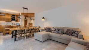 Un salon moderne comprenant un canapé d’angle, une table à manger avec des chaises et un coin cuisine doté de meubles en bois et d’un éclairage suspendu.