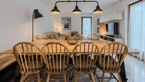 Un coin repas avec une table et des chaises en bois, un luminaire suspendu noir, un canapé, un tableau et un téléviseur posé sur un meuble.