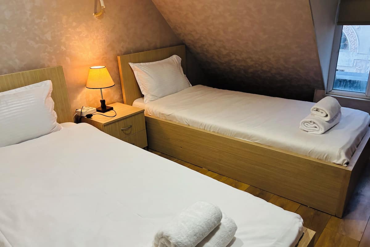 A hotel room with two beds, a nightstand with a lamp, and a window.