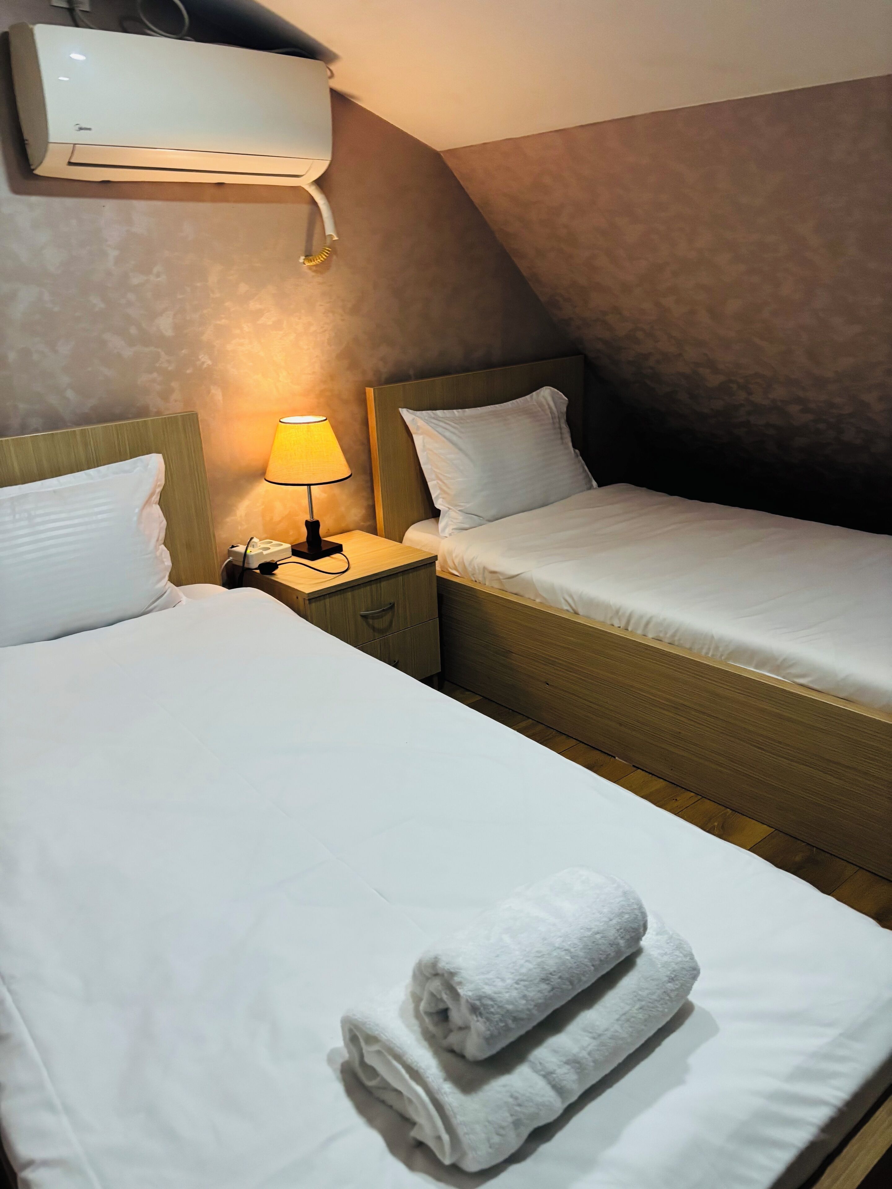 A hotel room with two beds, a bedside lamp, a wall-mounted air conditioner, and a wooden headboard.