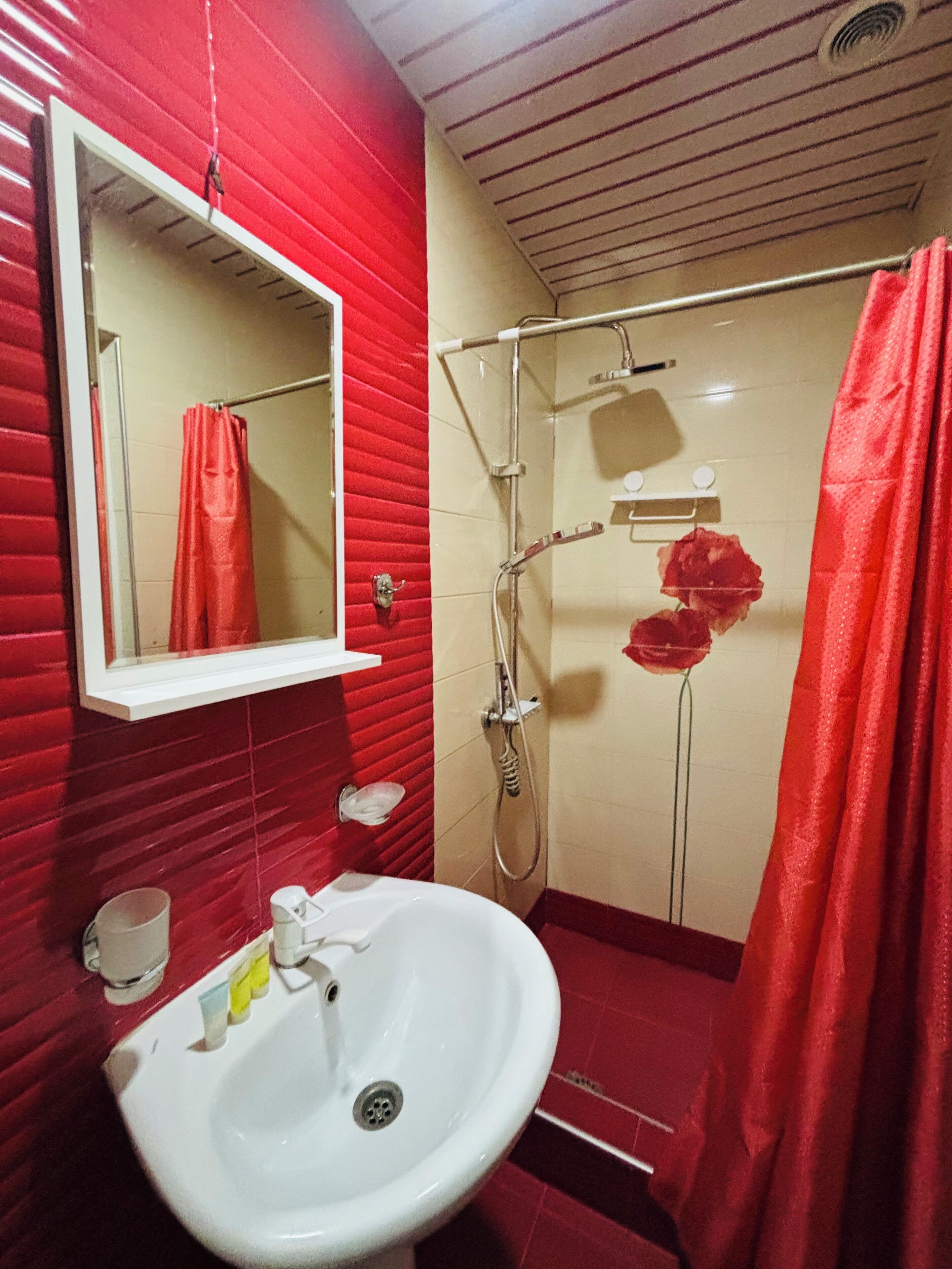 A bathroom with a red tiled wall, a white sink, a shower with a red shower curtain, and a mirror above the sink.