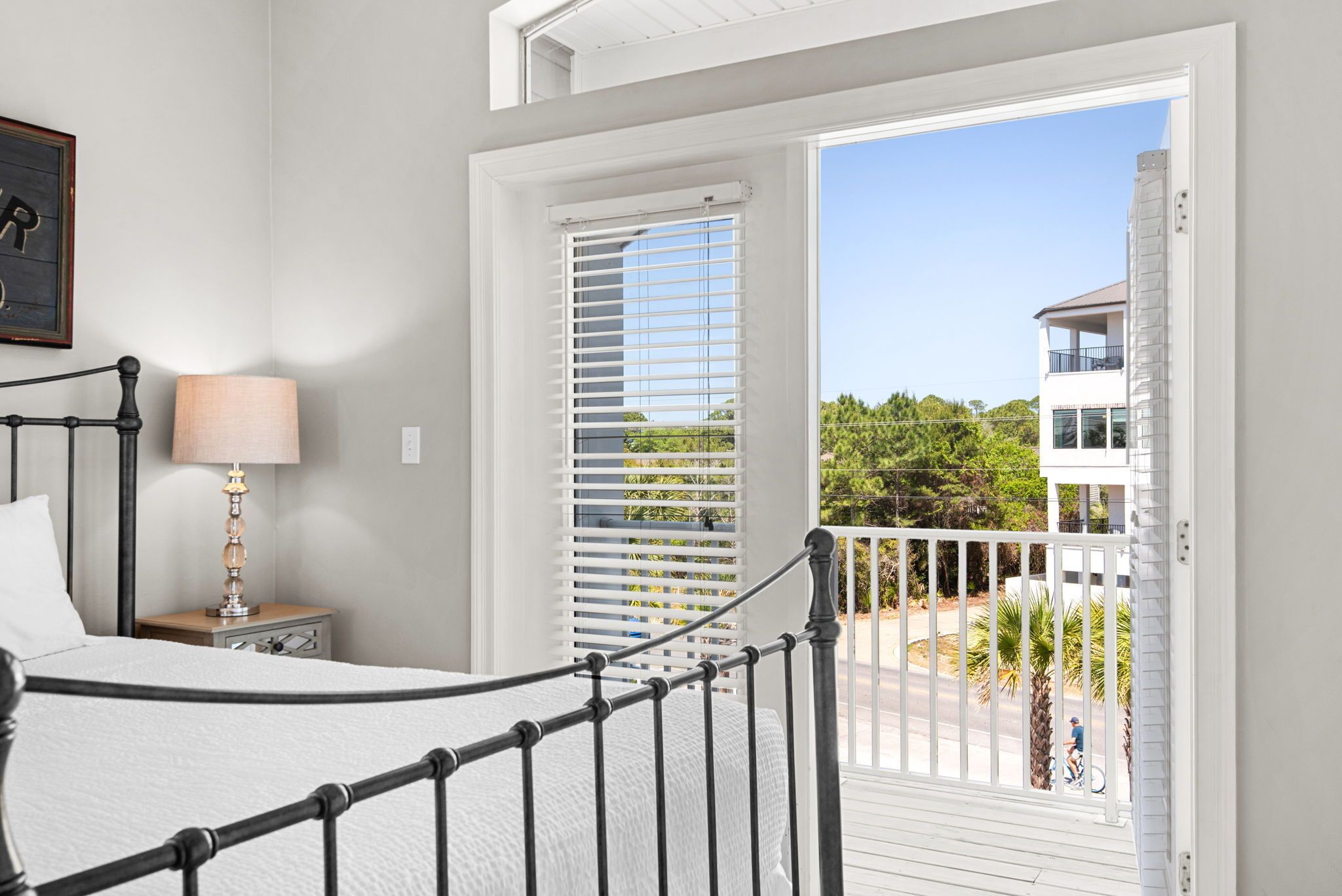 A bedroom with a bed, bedside table, lamp, and a view of a balcony with a railing and greenery.