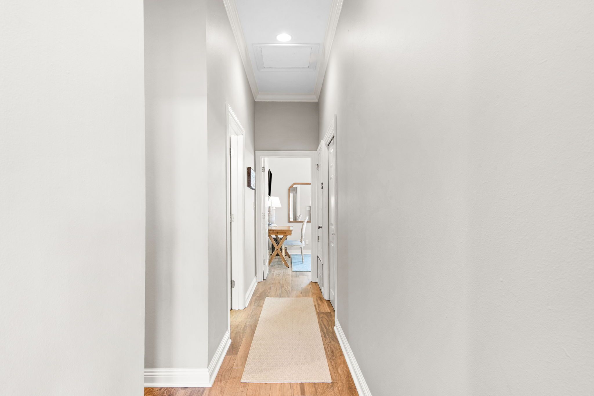 Un couloir doté d’un plancher en bois, de murs blancs et d’une porte au fond, qui donne sur une pièce contenant une chaise et une table.