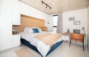 Quarto com cama, escrivaninha, cadeira e janela com cortinas.