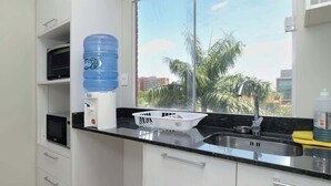 Una cucina moderna con mobili bianchi, un piano di lavoro nero, un dispenser d'acqua, un lavello e una finestra con vista su palme.