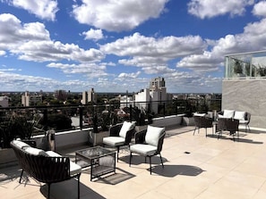 Eine Dachterrasse mit Außenmöbeln, einer Glasbrüstung und Blick auf die Stadt.