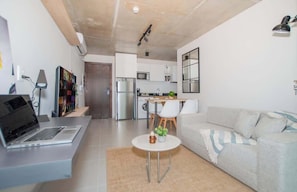 A modern living room with a sofa, a small round table with a plant, a laptop on a desk, and a kitchen in the background. - Modern Apt w/City View | Gym, Pool | Walk to Malls (Asunción)