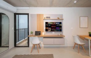 Una sala de estar moderna con una puerta de vidrio, un escritorio de madera con una computadora portátil, dos sillas blancas y un estante montado en la pared con diversos objetos.